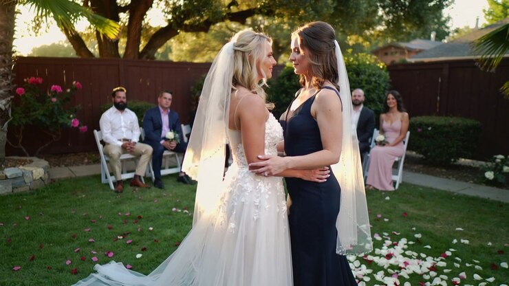 Embracing, two brides standing in backyard at wedding while camera moving closer, gowns and petals