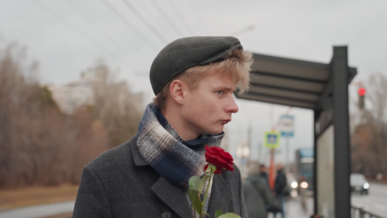 Giovane creatore caucasico che offre una rosa con un'aria da flirt urbano giocoso e sicuro di sé, con sciarpa e berretto, sguardo audace, primo piano della rosa vicino alle labbra, luce nuvolosa, pensilina del tram e tram sullo sfondo, performance affascinante.