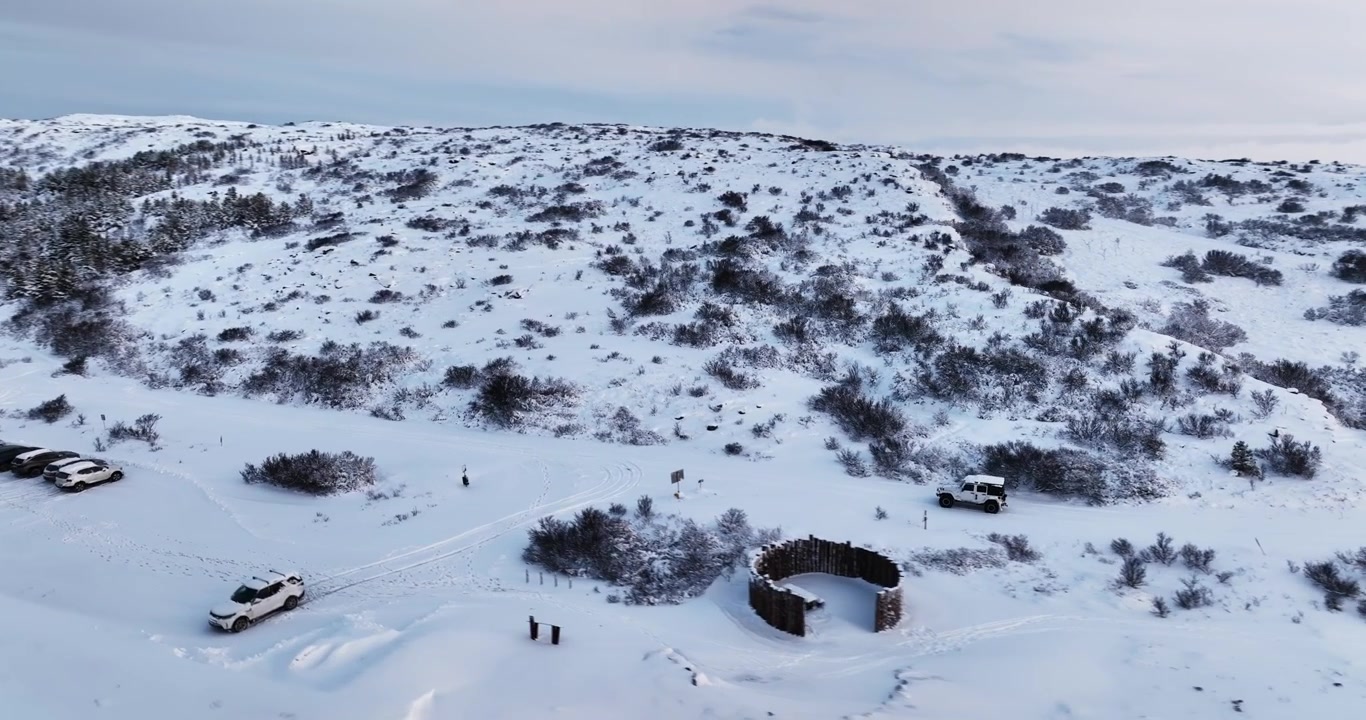 Drone view of a white 4x4 in snowy Heiðmörk, Iceland, near small shelters and volcanic winter hills with dark shrubs, tire tracks, and open frozen terrain.
