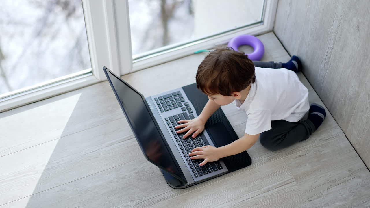 A little boy sits on the floor and plays on a laptop. The child is sitting on the floor near the window and looks at the screen.