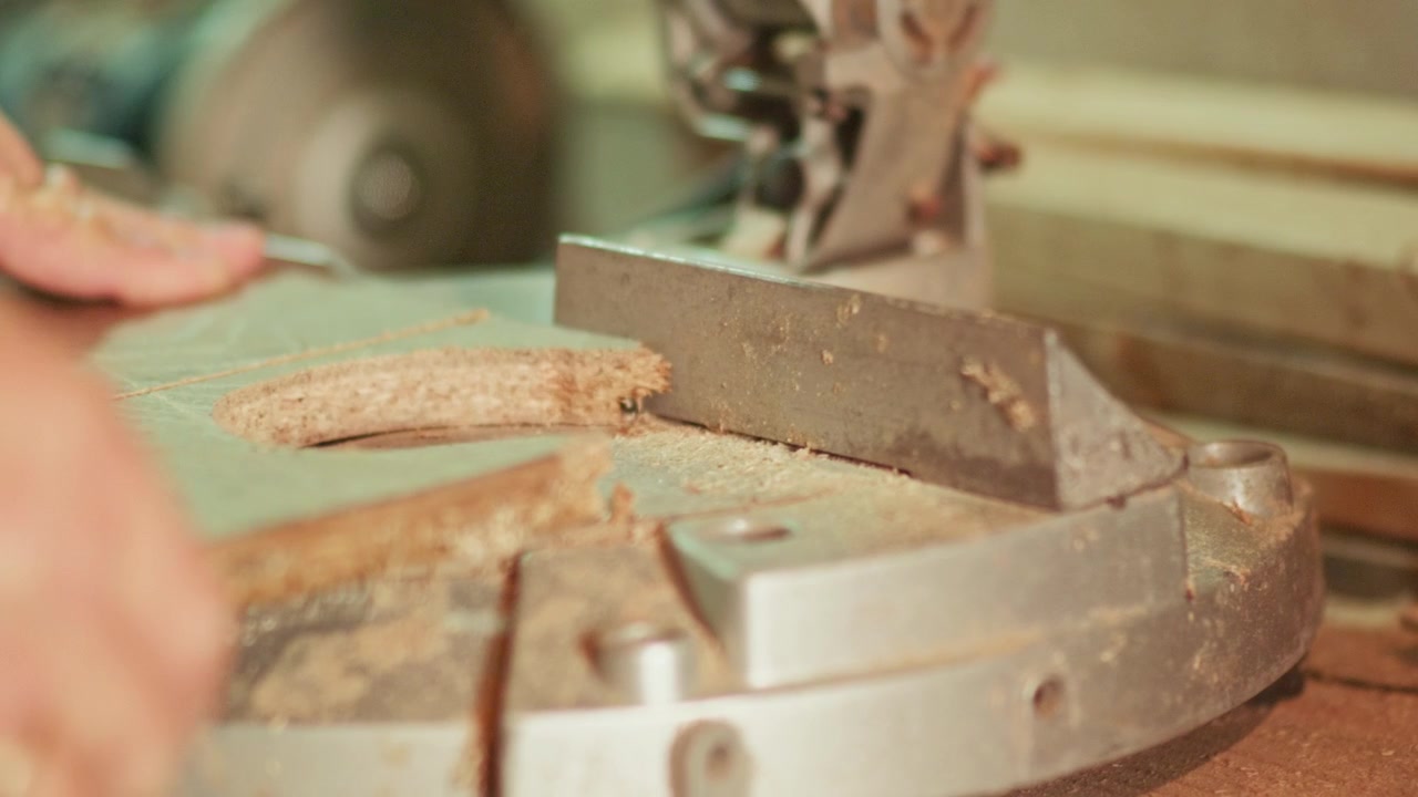 Nahaufnahme eines Handwerkers, der mit einer elektrischen Kappsäge ein breites Holzbrett in einer Werkstatt zuschneidet. Fliegende Sägespäne und präzise Klingenführung für Tischler- und Bauprojekte
