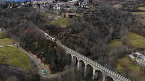 Antena:-Tren-De-Pasajeros-Cruzando-Un-Puente-De-Piedra-En-Un-Paisaje-Montañoso,-Ciudad-Montañosa-Y-Picos-Nevados-En-El-Fondo