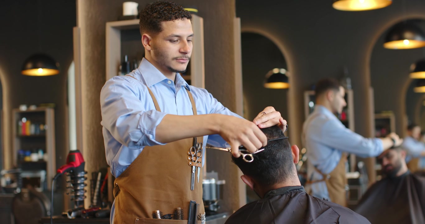 Afroamerikansk barber sætter en klemme, klipper hår med kam i salonen iført læderforklæde til formgivning.