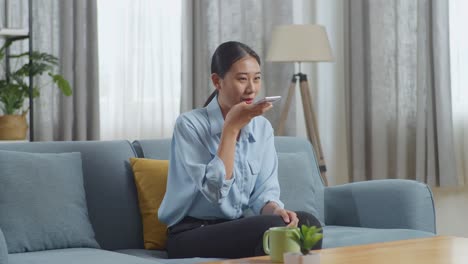 asian woman recording voice message on smartphone while sitting on sofa in the living room