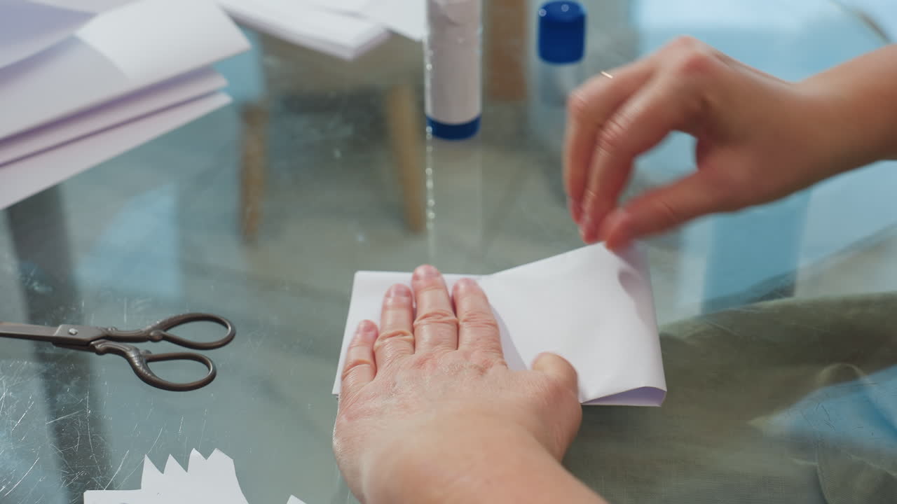Hand view of woman creating paper craft, ensuring the edges are equal, scissors and liquid glue visible on table, emphasizing attention to detail in crafting process with a calm home setting