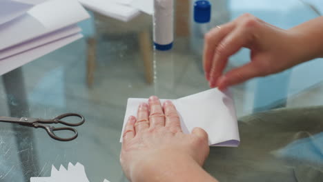 hand view of woman creating paper craft, ensuring the edges are equal, scissors and liquid glue visible on table, emphasizing attention to detail in crafting process with a calm home setting