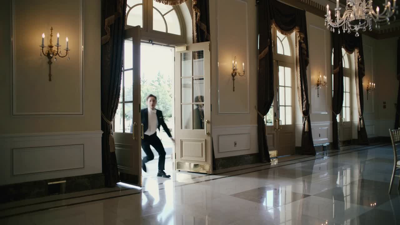 Rushing man in tuxedo walking through open double doors into ballroom, scanning for missing guests