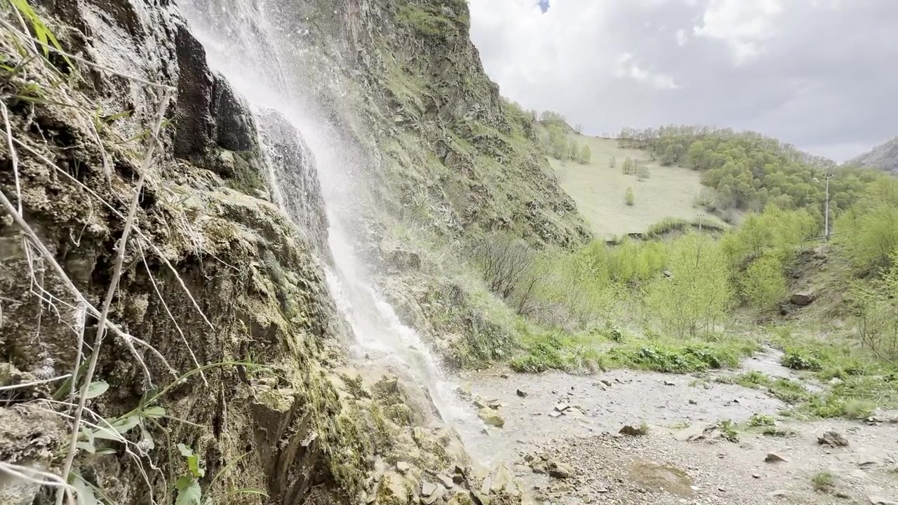 Una piccola cascata scende da una parete rocciosa nel Distretto di Malokarachayevsky, Karachay-Circassia, Russia, con lussureggianti montagne verdi sullo sfondo sotto un cielo nuvoloso.
