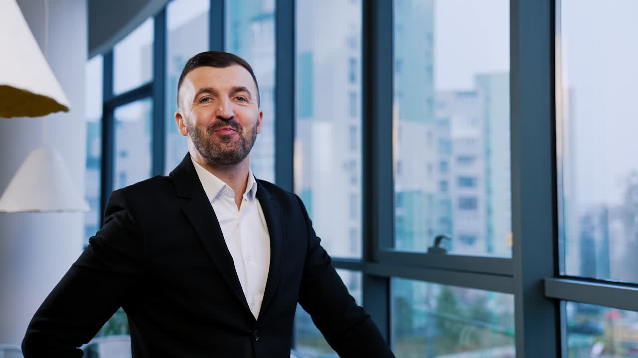 Good-looking bearded man stands at window and turns to camera. Businessman looking straight into camera smiling. Cityscape in blur at backdrop.
