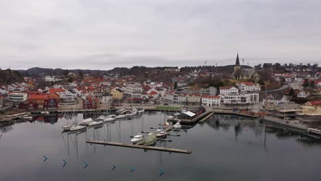 Puerto-Deportivo-Y-Centro-De-La-Ciudad-De-Grimstad---Hermosa-Antena-Matutina-Moviéndose-Lentamente-Hacia-Atrás-Sobre-La-Superficie-Clara-Del-Mar-Para-Revelar-Toda-La-Ciudad-Cerca-De-La-Carretera-Arendal---Noruega