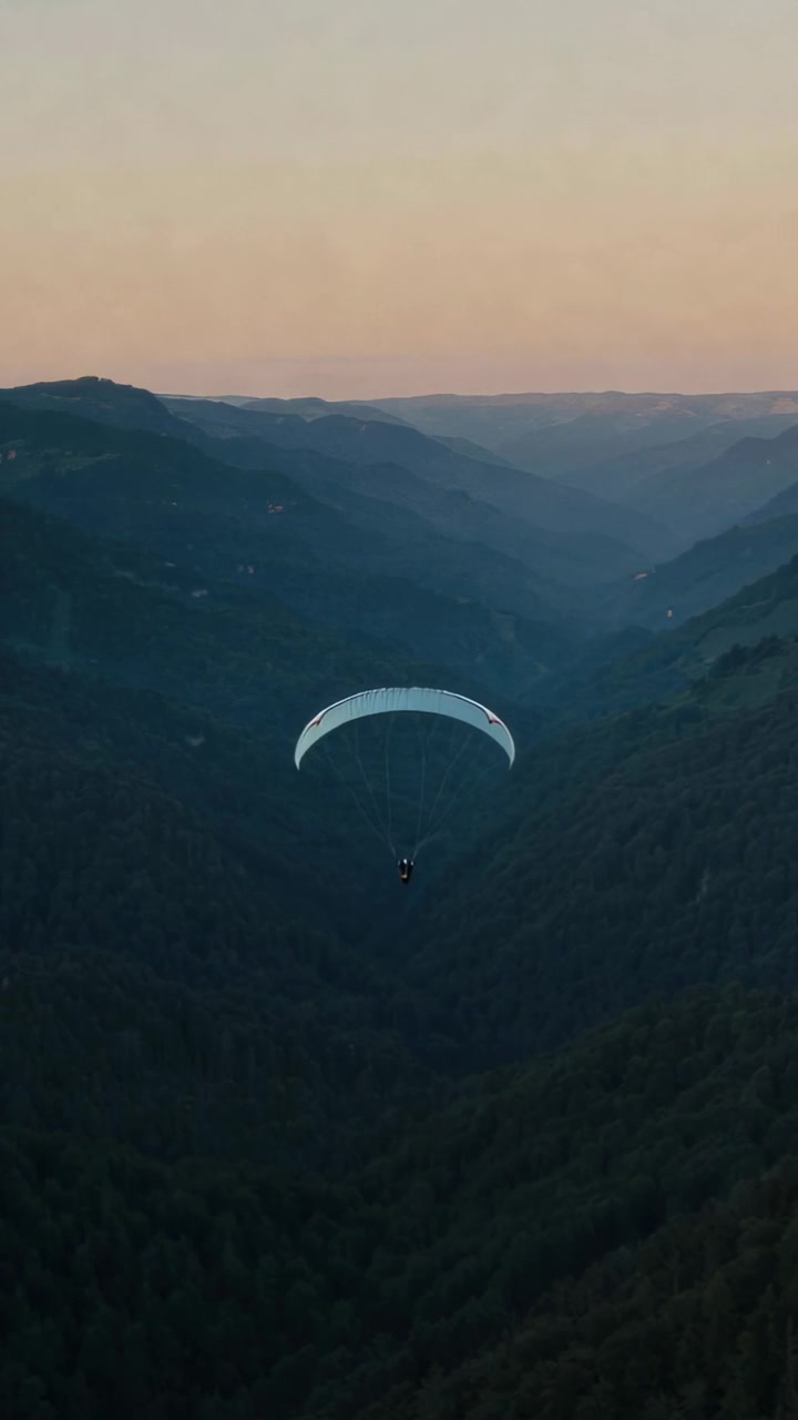 Pilota che regola i controlli mentre naviga con una vela da parapendio giù per una profonda valle tra creste che sfumano al tramonto.