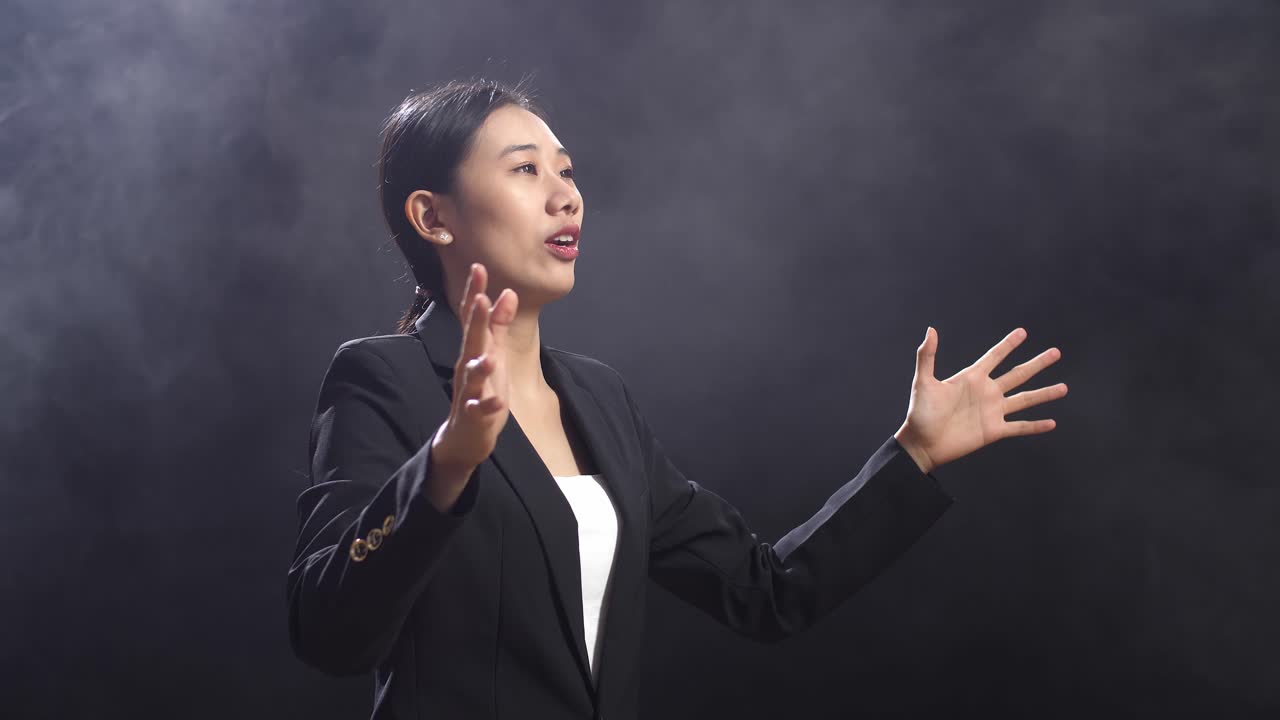 블랙 스크린 스튜디오 에서 연설 하는 동안 손 을 고 있는 비즈니스 슈트 를 입은 아시아어 연설자 여자 의 측면 시각