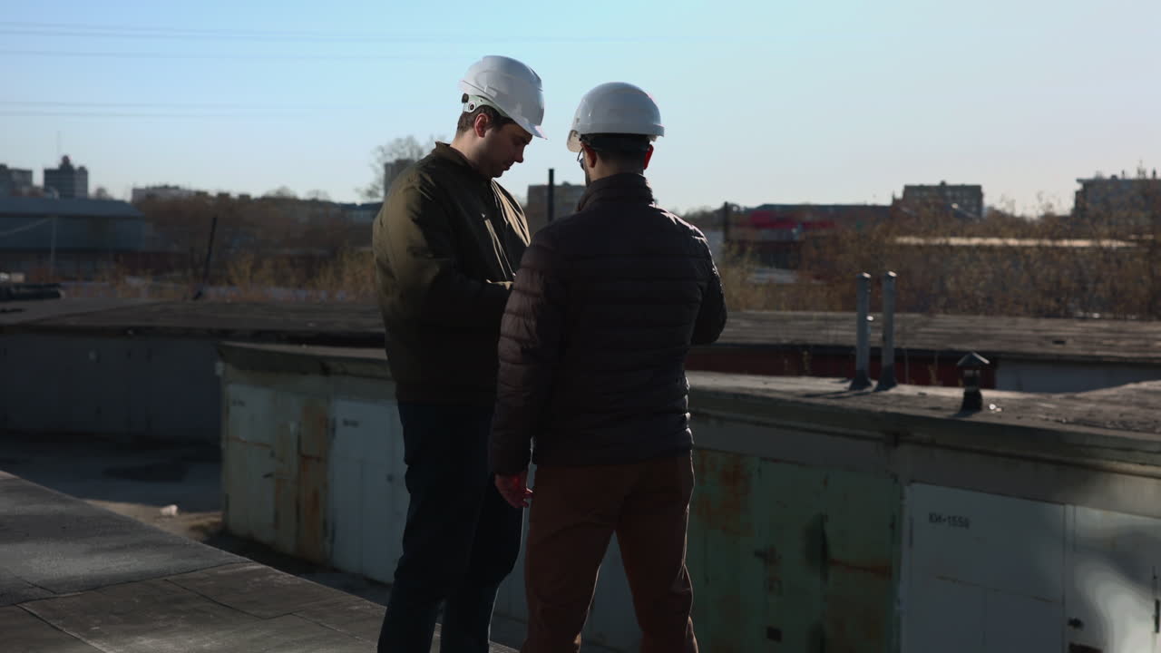 Two Engineers On Rooftop Review Plans On Tablet While Discussing Structural Details And Next Steps