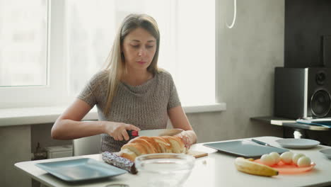 medium shot of young woman seated by window slicing loaf of bread into equal pieces on wooden board next to eggs banana and plates under natural morning light streaming through glass