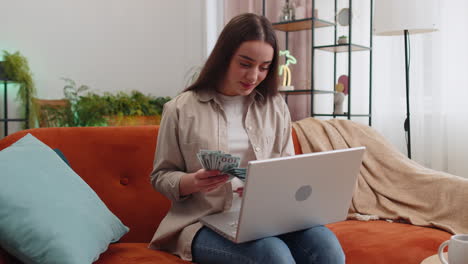 Una-Mujer-Feliz-Y-Sonriente-Contando-Dinero-En-Efectivo-Y-Usando-Una-Computadora-Portátil-Para-Calcular-Los-Ingresos-Domésticos-En-Casa