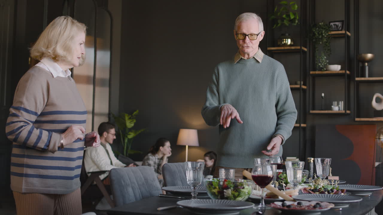 abuelos felices preparando la mesa del comedor, mientras que en el fondo su familia se sienta en la sala de estar y habla juntos
