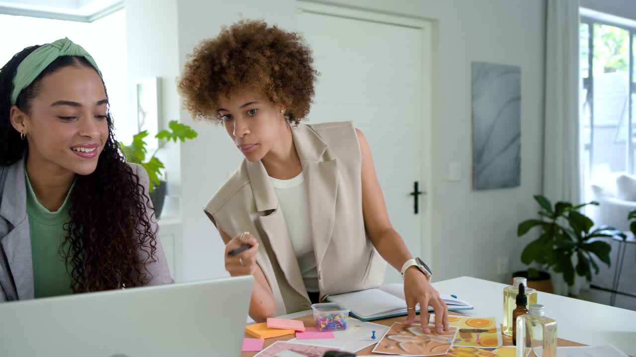 Wanita Afrika-Amerika mendesain di meja, ada yang berdiri menunjuk contoh warna, ada yang duduk mengetik dan mengedit di laptop.