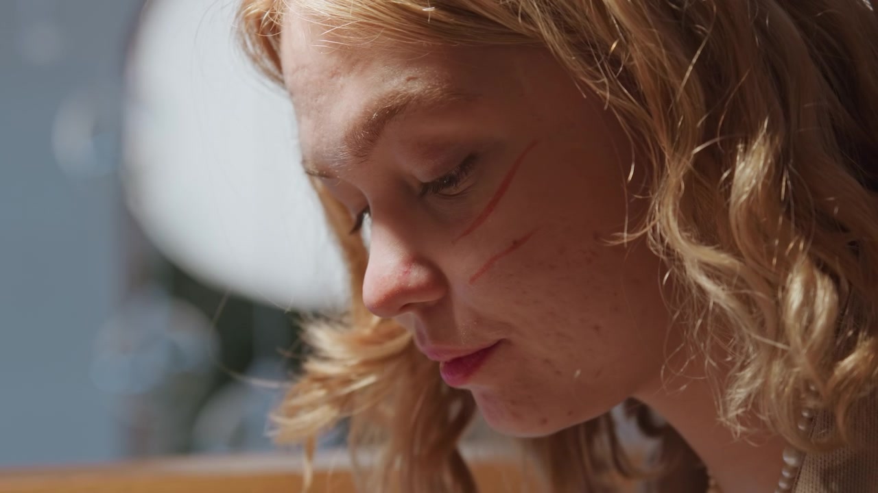 Close-up de jovem mulher com cabelo loiro encaracolado, vestindo roupa de cor clara e colar de pérolas, sob iluminação interna suave. A mulher está focada em comer de um prato em uma mesa de madeira perto da janela.