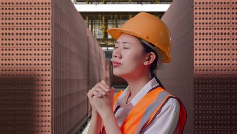 close up side view of asian female engineer with safety helmet pray for something while standing with red brick packed in stacks are stored