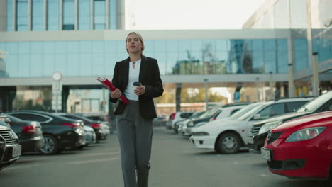 confident young businesswoman in formal outfit holding folder drinking coffee walking through parking lot with modern glass building background during busy workday morning commute