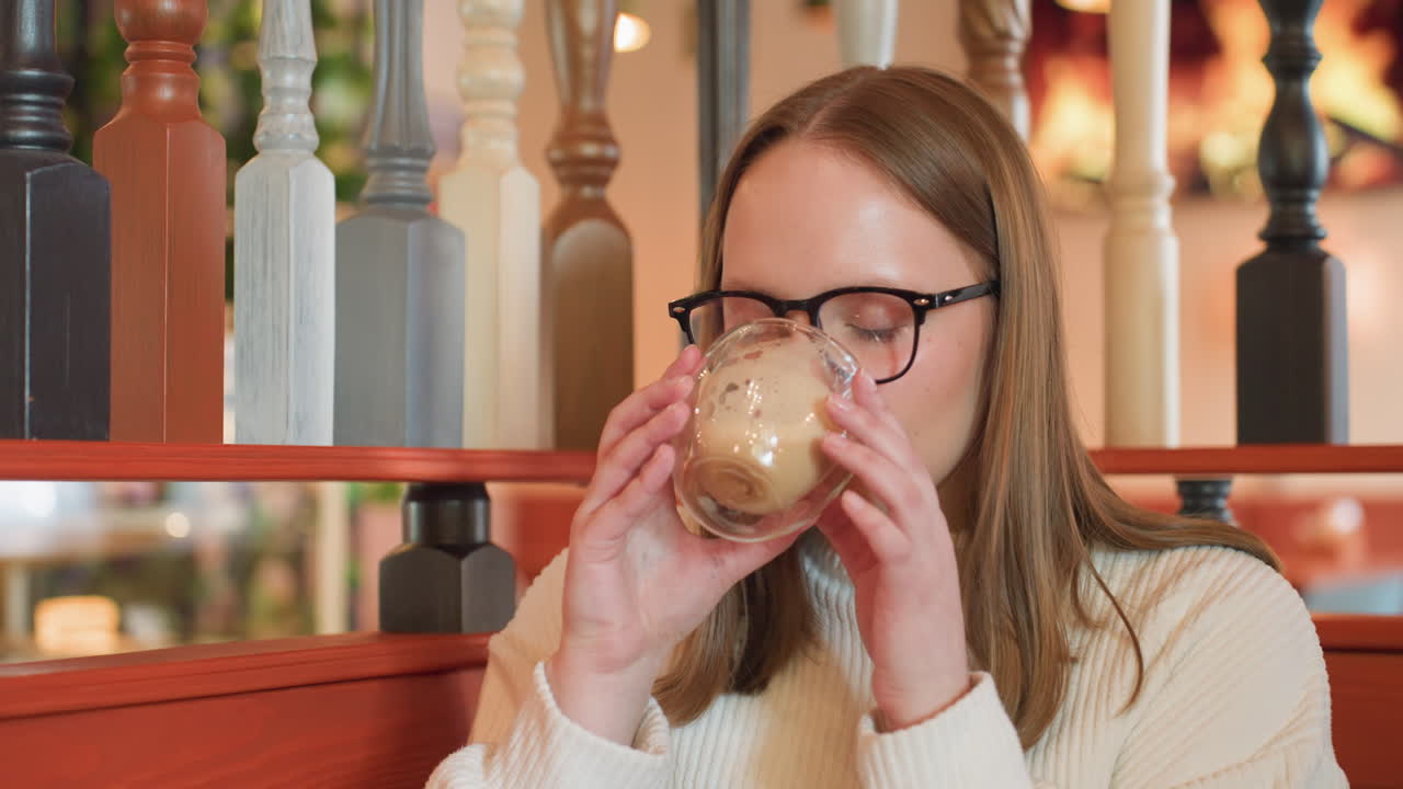 young woman in white sweater sitting inside cozy cafe, wearing glasses and looking down with calm expression, soft background with wooden railing