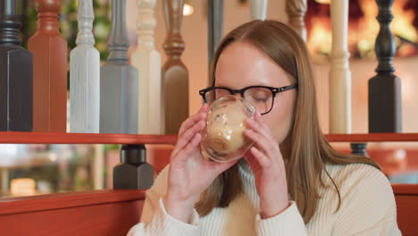 young woman in white sweater sitting inside cozy cafe, wearing glasses and looking down with calm expression, soft background with wooden railing