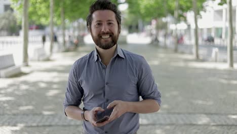 hombre barbudo usando un teléfono inteligente y sonriendo a la cámara