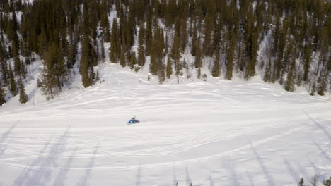 Speedy-snowmobile-travelling-snowy-arctic-circle-alpine-woodland-trail-aerial-view-above-Lapland-forest