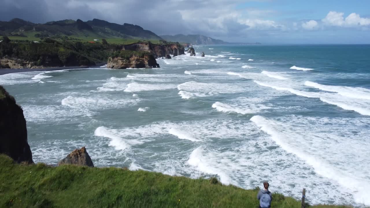 Drone view of a person standing on a cliff and the coast, rocks and ocean on a sunny summer day at Three Sisters and Elephant Rock, New Zealand