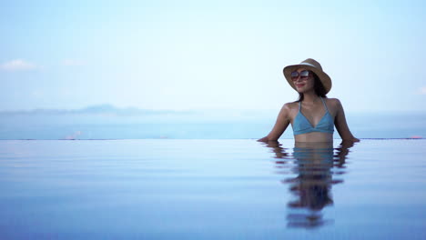 Eine-Hübsche-Dame-In-Einem-Blauen-Badeanzug-Mit-Sonnenhut-Und-Sonnenbrille,-Die-Allein-In-Einem-Friedlichen-Infinity-Pool-Steht-Und-Ihr-Spiegelbild-Auf-Ruhigem,-Klarem,-Blauem-Wasser-Scheint