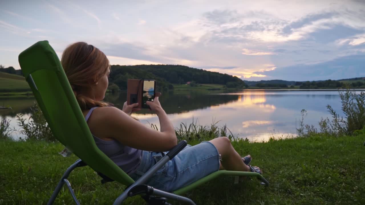 Donna seduta su una sedia da esterno che fotografa un lago calmo e un cielo al tramonto con lo smartphone, catturando uno stile di vita da campeggio tranquillo e un paesaggio naturale suggestivo a Savci, Slovenia