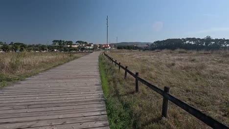 First-person-view-down-the-wooden-path-in-sandy-terrain-of-dry-vegetation-towards-the-buildings-on-a-bright-and-sunny-summer-day-on-vacation,-shot-traveling-forward