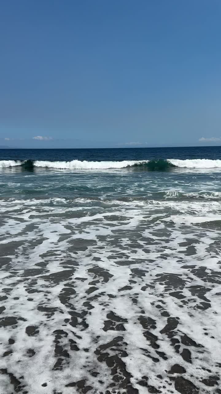 Onde dell'oceano che si infrangono su una spiaggia di sabbia scura sotto un cielo azzurro e limpido