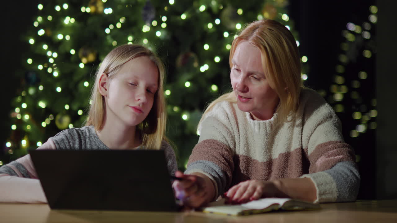 Mom and daughter are looking at the laptop screen together. Sitting in front of the blurred lights of the Christmas tree