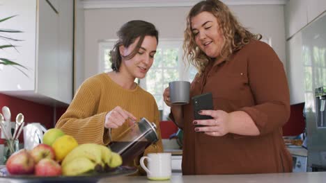 feliz pareja de lesbianas caucásicas vertiendo café y usando teléfono inteligente en la cocina