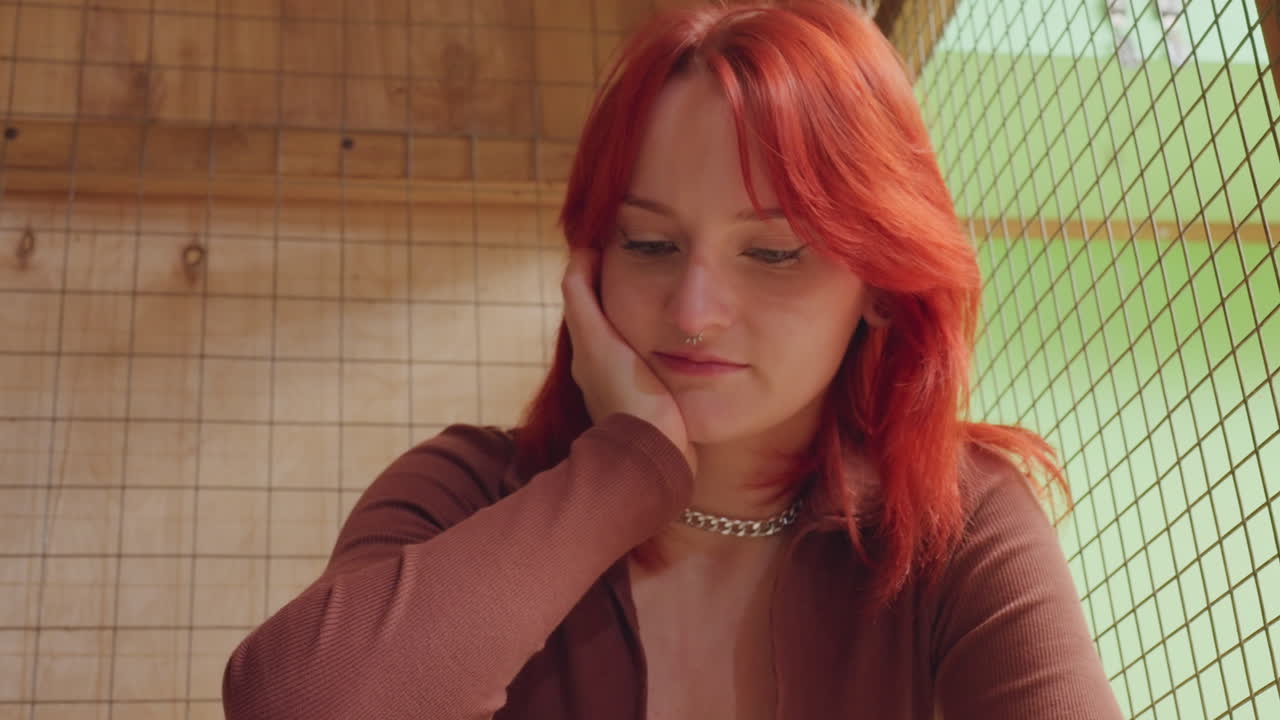 Donna caucasica dai capelli rossi seduta con un gatto, ambiente rustico con sfondo a rete verde, espressione pensierosa, collana a catena, maglione marrone, mano delicata sulla pelliccia, illuminazione calda e soffusa, ritratto intimo