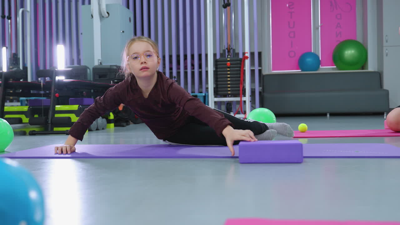 joven en una alfombra de gimnasio colocando un bloque púrpura en la cintura, las piernas de otro niño visibles, rodeada de varios equipos de gimnasia en un entorno colorido