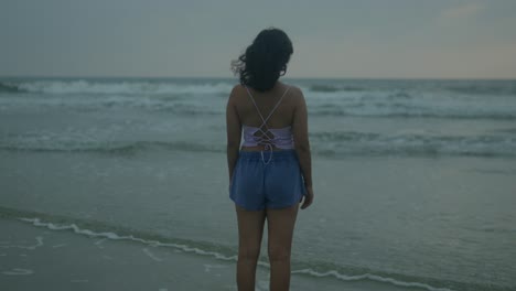 a woman in a strappy top and denim shorts stands at the edge of the ocean, gazing at the twilight waves