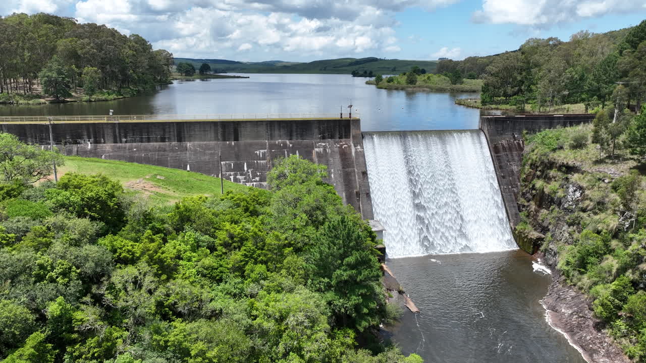 Continuation of previous shot. Drone flies close to the waterfall formed by Barragem do Divisa, then ascends to show the dam and surrounding forested landscape in São Francisco de Paula, RS