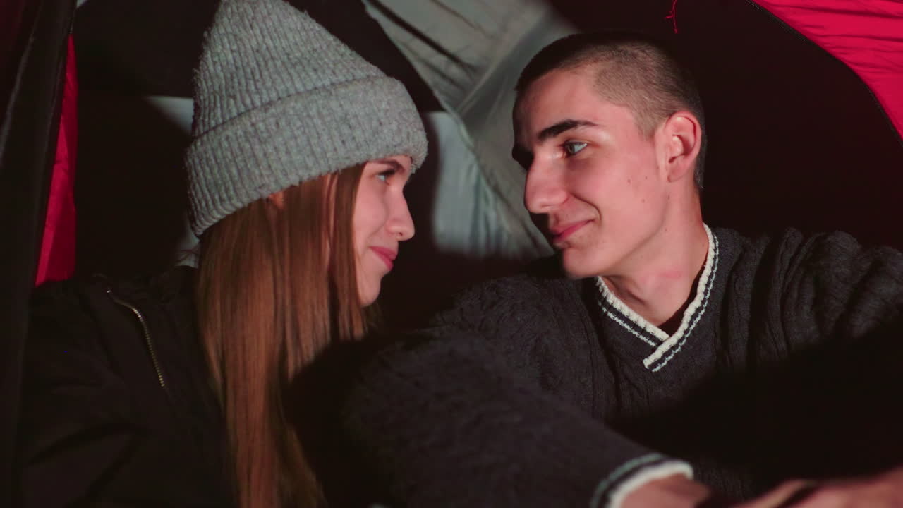 Young couple sitting close inside tent at night exchanging heartfelt smiles as woman keeps smiling warmly while man smile slowly fades, capturing tender emotional shift in quiet moment
