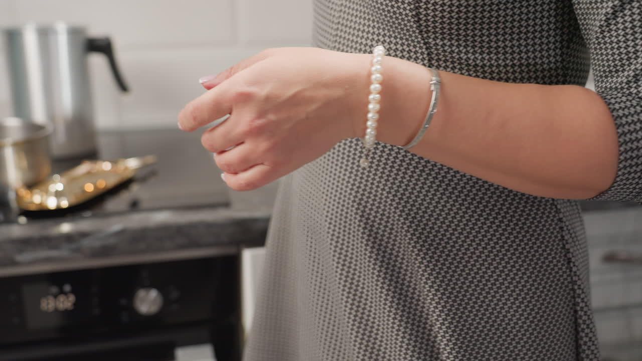 Culinary artist folds kitchen towel with both hands, wearing elegant bead bracelet and silver bangle, preparing to toss towel onto gas stove in clean modern kitchen with stainless cookware
