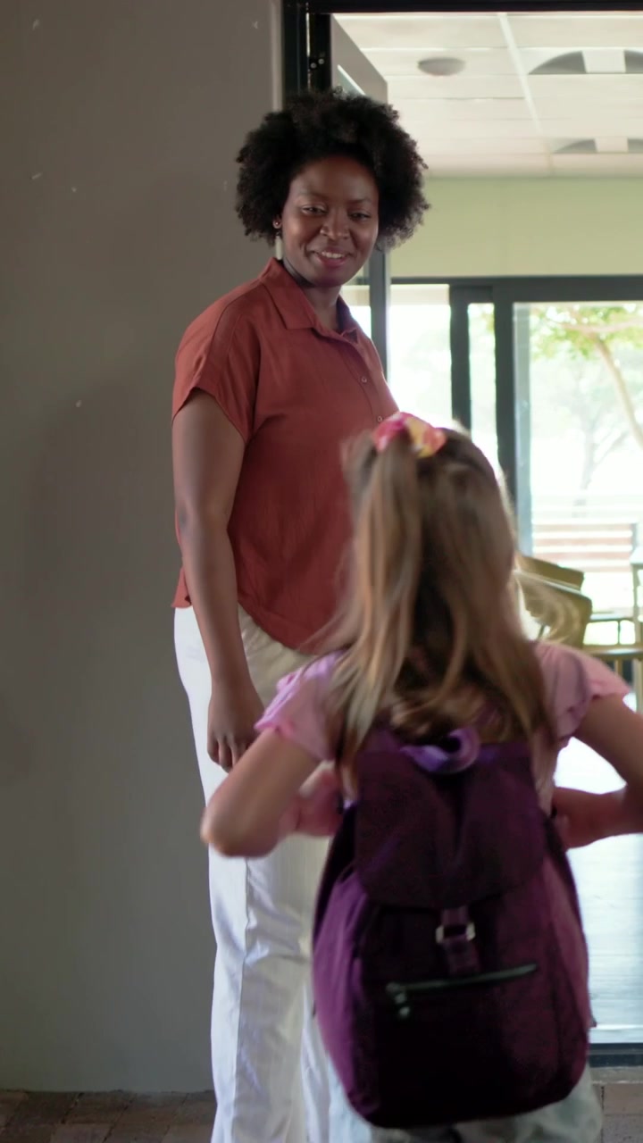 Vertical video: Welcoming woman in rust shirt stepping aside at door, taking backpacks as girls run