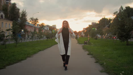 sick girl covering herself with coat strolling along paved path with chair behind and blurred view of people moving in busy city during sunset with green trees grass roadside buildings and cloudy sky