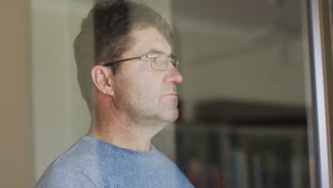 hombre caucásico pensativo con gafas mirando por la ventana en casa