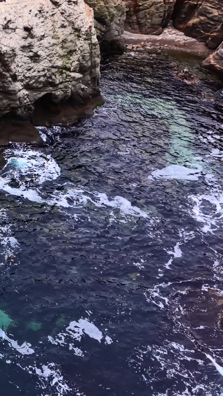 Vista dall'alto di acqua oceanica blu scuro che colpisce la base di scogliere di roccia grigia in una cala. Acqua di mare tra ripide coste rocciose. Video verticale