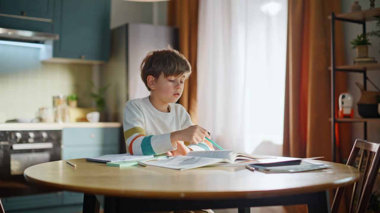 Fatigued schoolboy reading textbook sitting kitchen. Tired primary school pupil