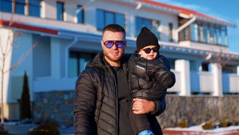 caucasian man wearing black jacket holds his little baby in hands. father and son wearing sunglasses stand outdoors on sunny day.