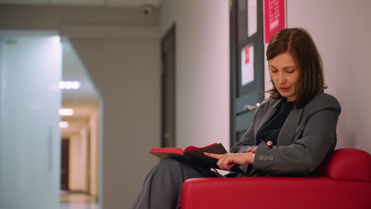 Businesswoman in gray suit sitting on red couch reading red notebook in office hallway. Calm, thoughtful expression with modern, professional environment visible in background