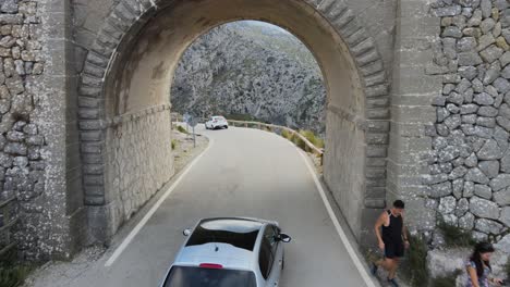 drone following a car driving under a bridge onto a winding mountain road in sa calobra, mallorca, spain