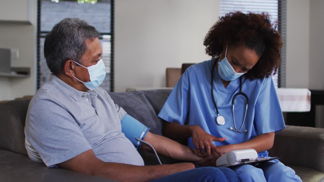 Mixed race female doctor wearing mask and senior man taking blood pressure at home